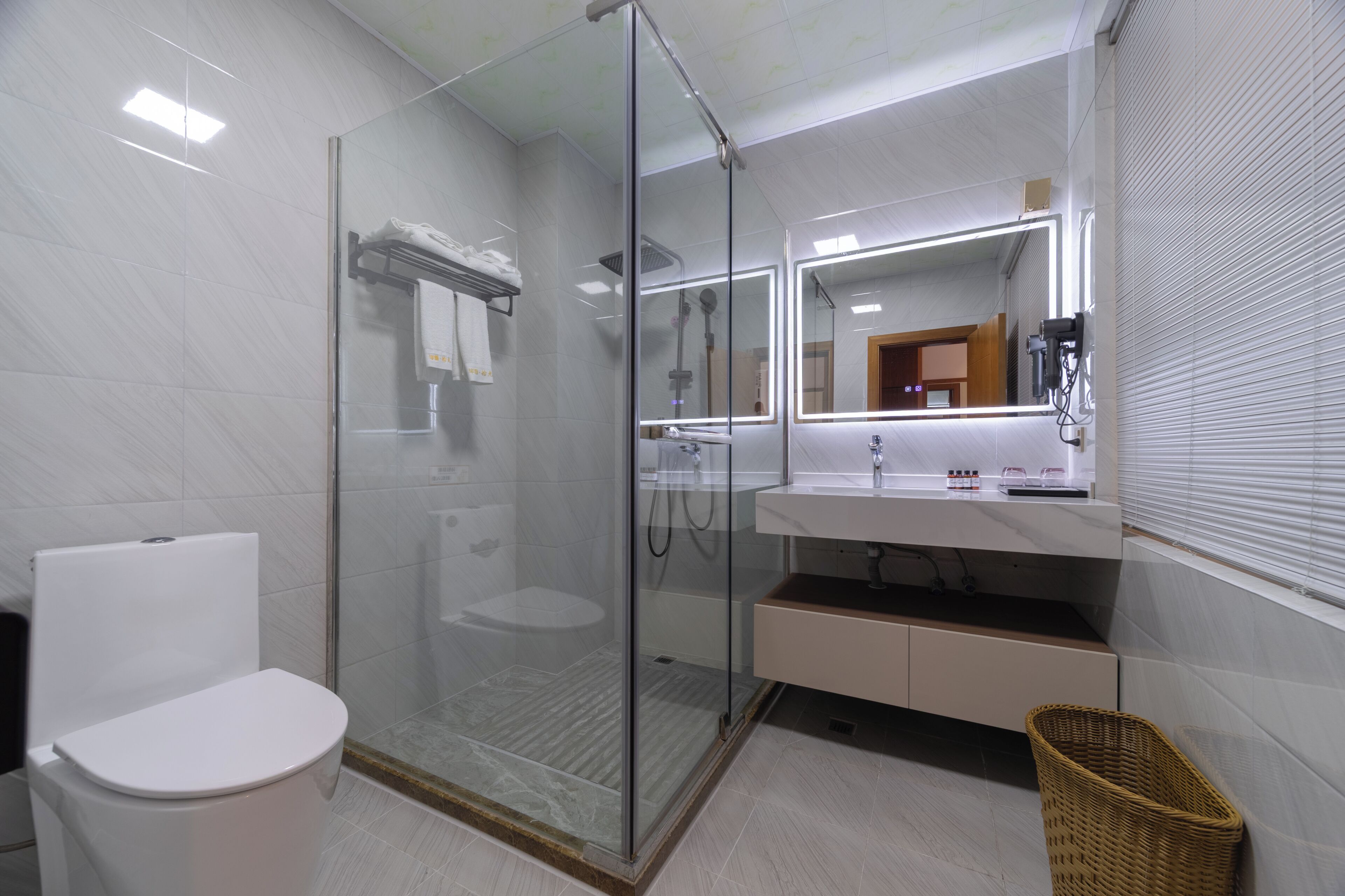 Un baño moderno con ducha envuelta en vidrio, inodoro blanco, tocador con espejo y cesta de mimbre.