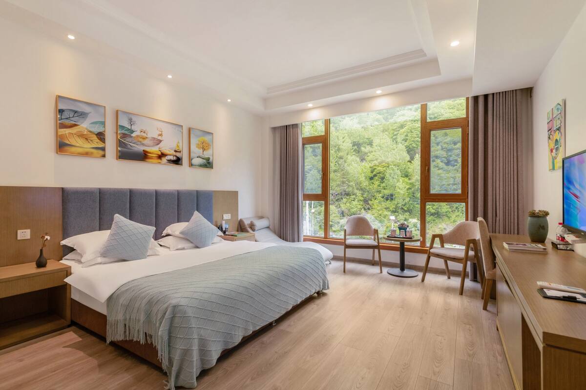 A hotel room with a large bed, a desk, two chairs, a TV, and a window with a forest view.