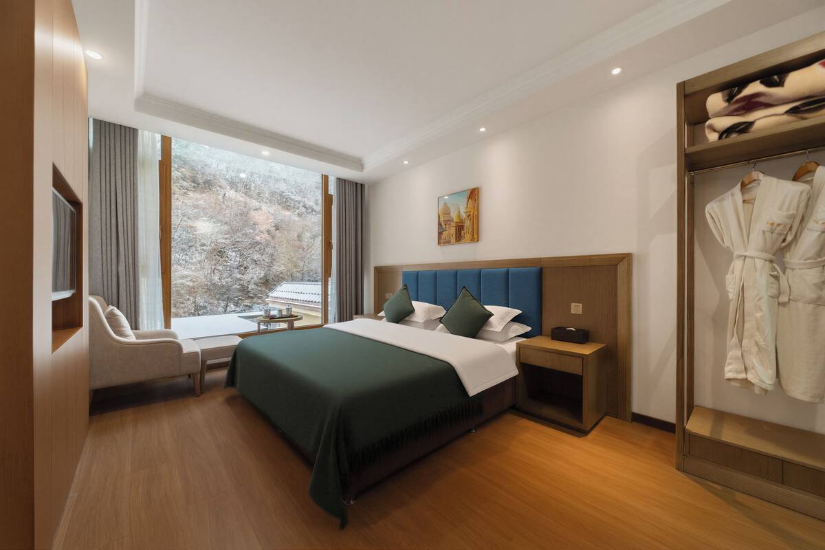 A hotel room with a bed, a seating area, a wardrobe, and a window with a view of trees.