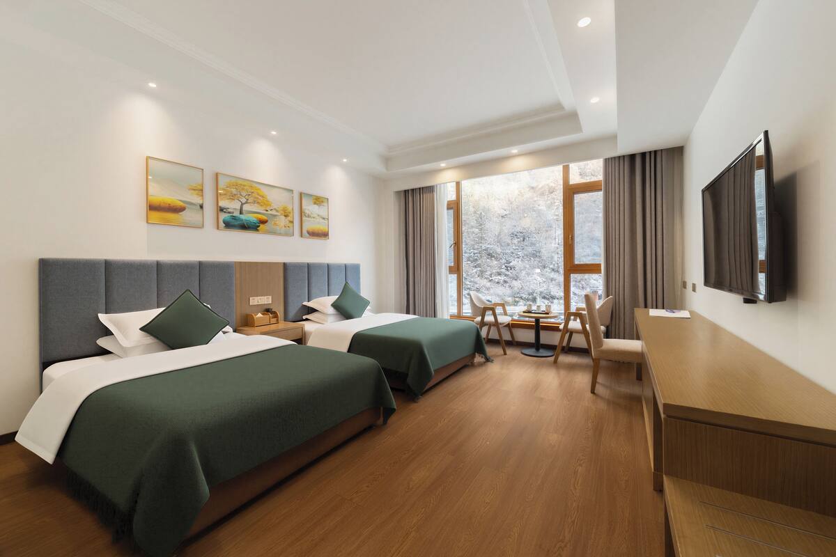 A hotel room with two beds, a desk, a chair, a TV, and a window with a view of trees.