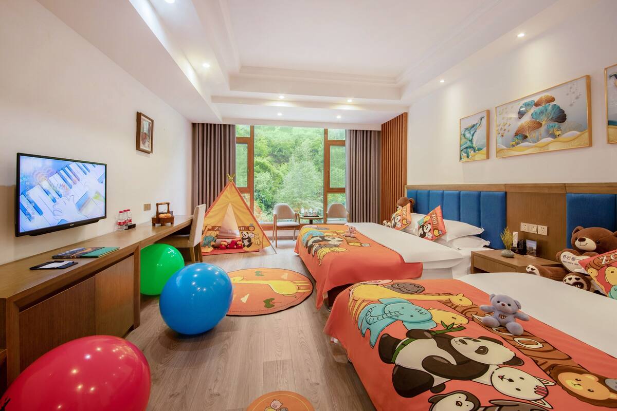 A hotel room with two beds, a TV, a desk, a chair, a tent, and a view of greenery outside.