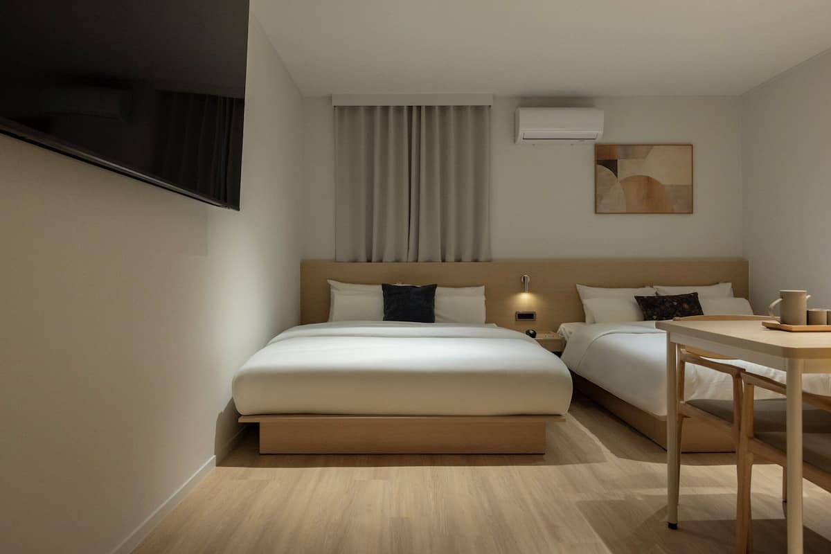 A hotel room with a bed, bedside tables, a desk, a TV, and an air conditioning unit.