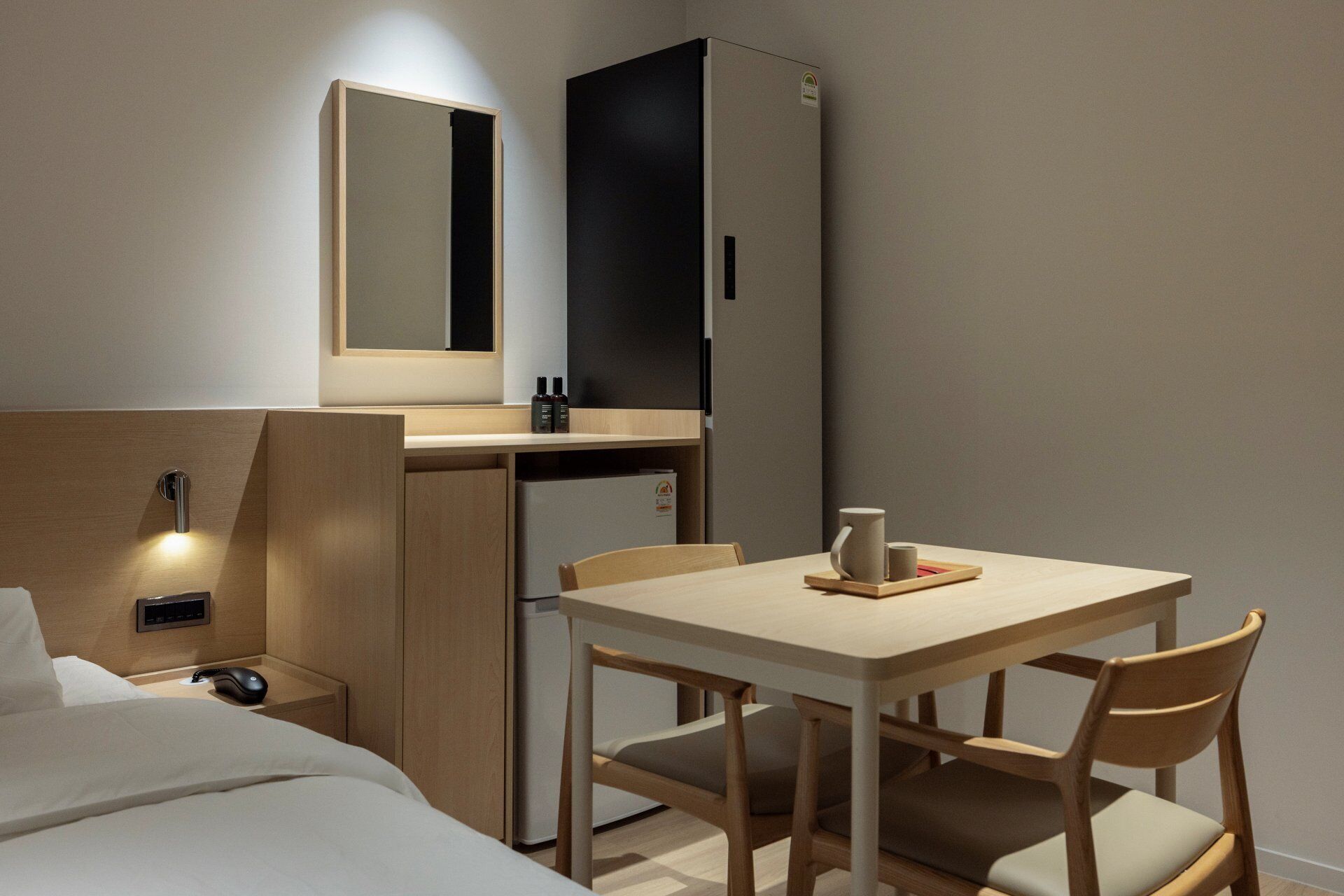 A hotel room with a bed, a desk with a chair, a small table with a cup, a mirror, and a nightstand with a phone and a lamp.