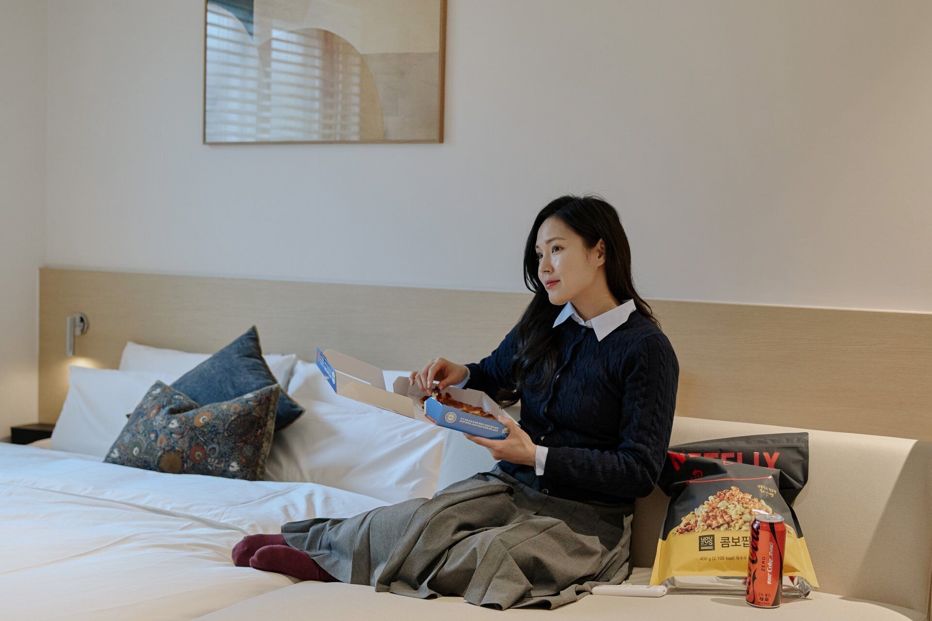 A person sitting on a bed with a pillow, holding a blue box, and a bag of popcorn on the bed.