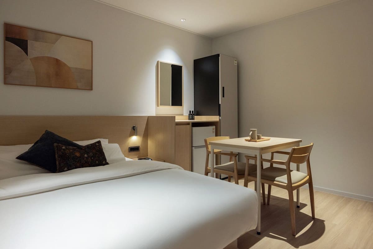 A hotel room with a bed, a desk with two chairs, a refrigerator, and a wall-mounted mirror.