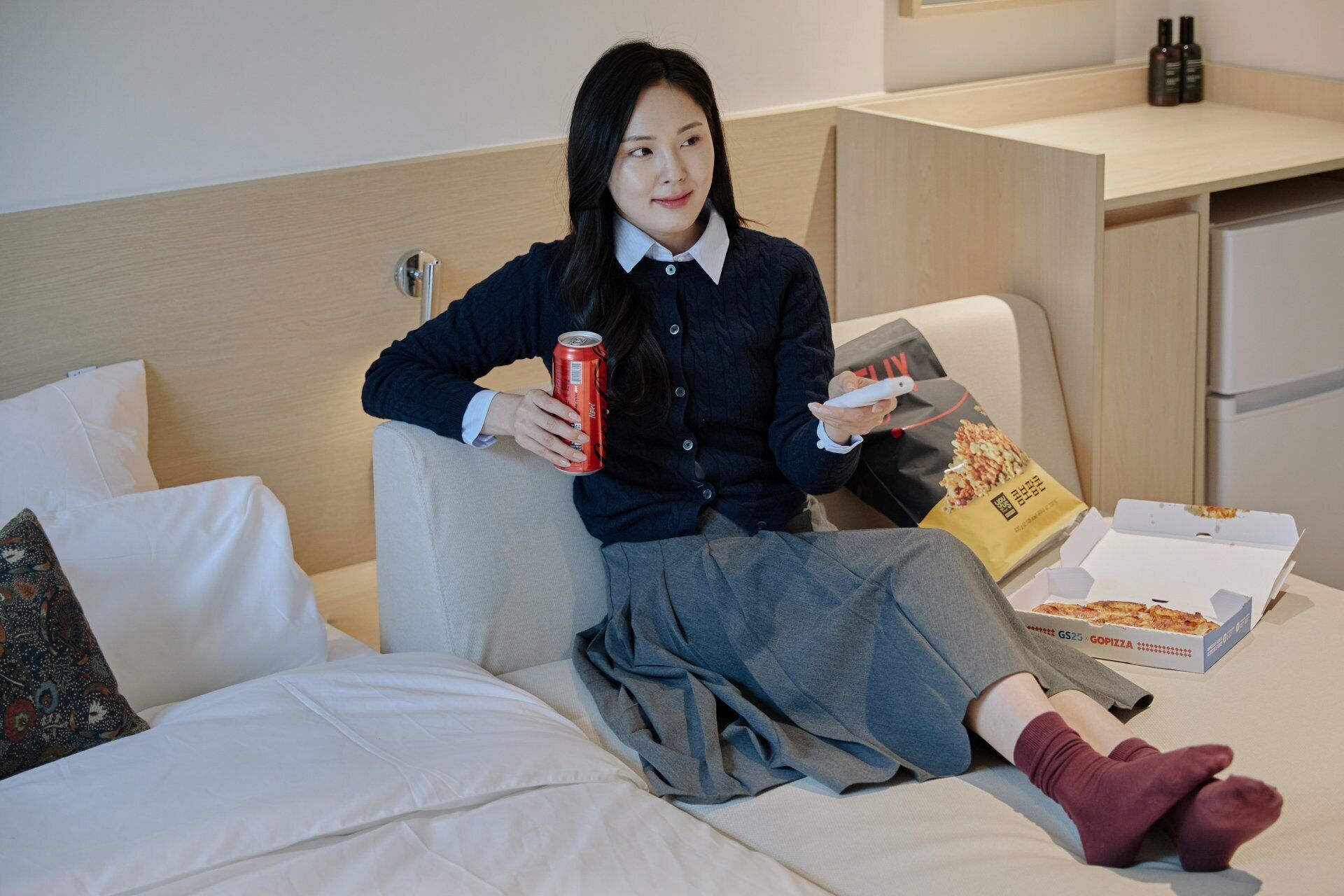 A person sitting on a bed with a pizza box, a can, and a pillow.