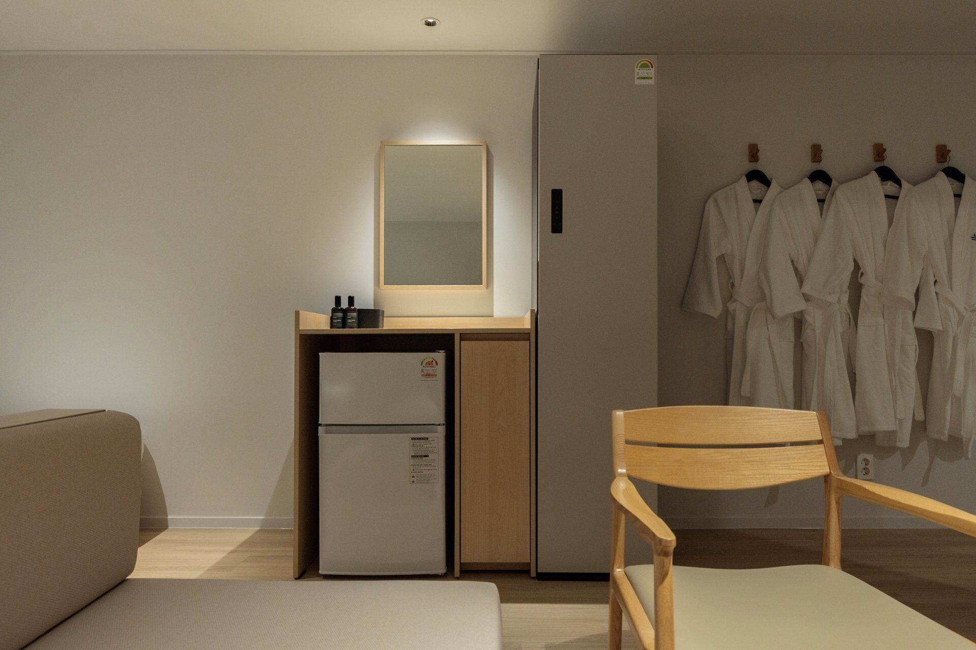 Habitación de hotel con cama, una silla de madera, un refrigerador pequeño, un espejo y un armario con batas.