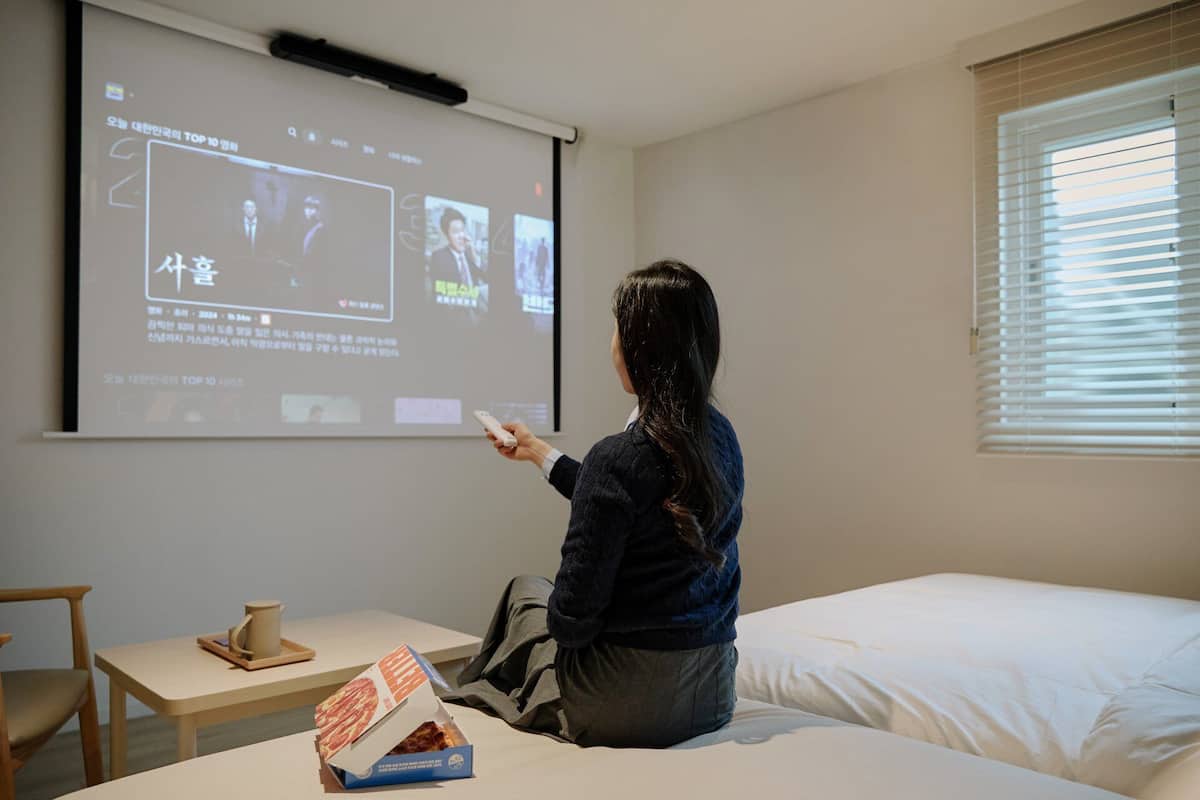 A person watching a presentation in a room with a bed, a small table, and a projector screen.