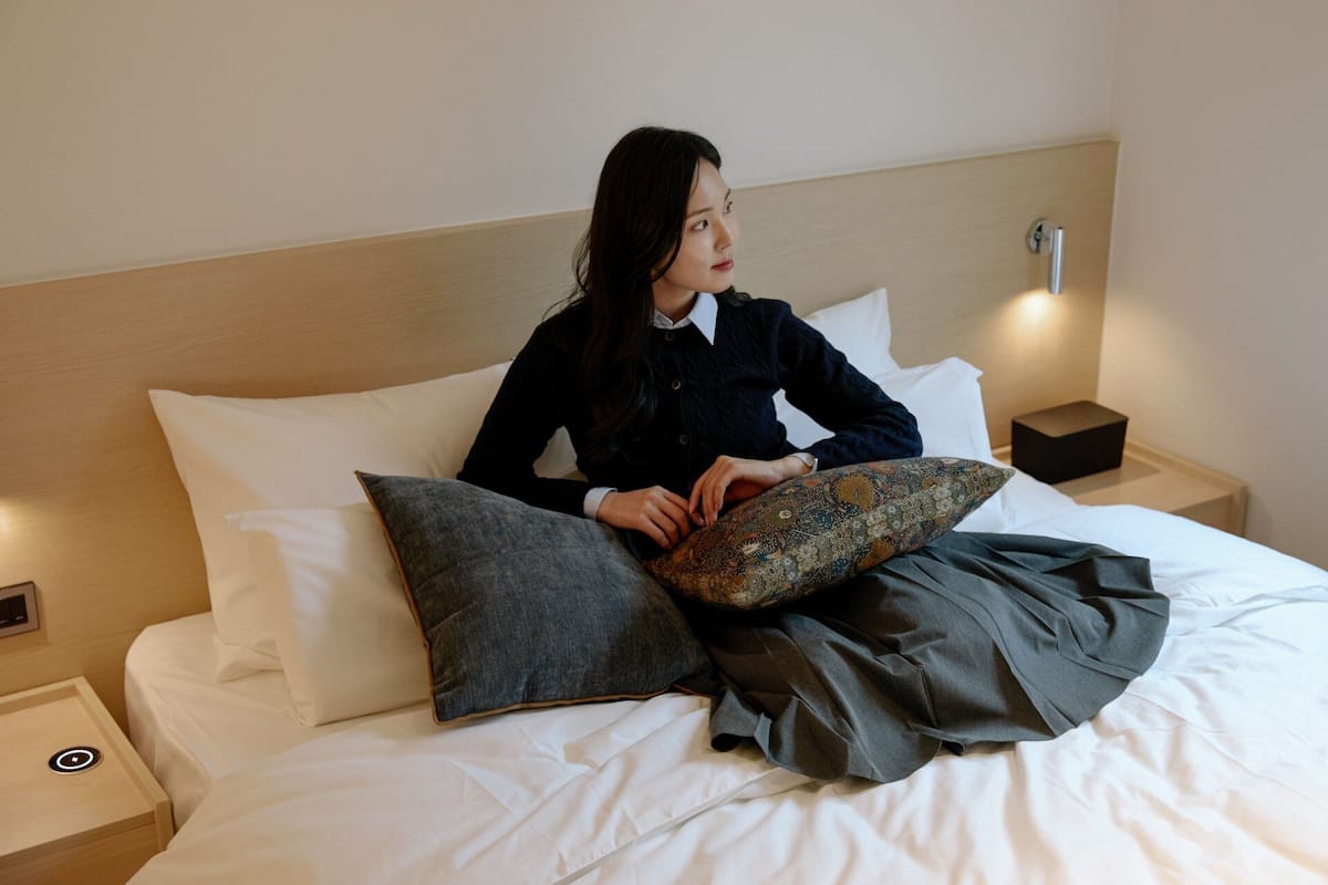 A person sitting on a bed with a wooden headboard, white bedding, and a small bedside table with a lamp.