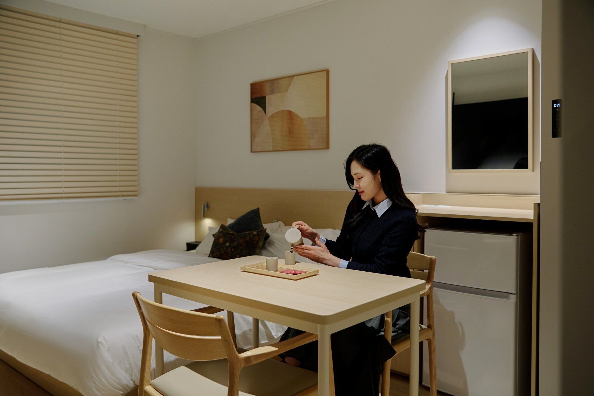 A hotel room with a bed, a table with chairs, a refrigerator, a wall-mounted mirror, and a painting.