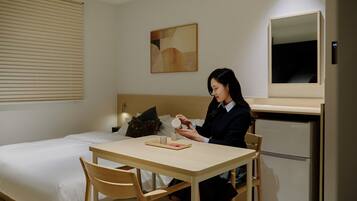 A hotel room with a bed, a table with chairs, a refrigerator, a wall-mounted mirror, and a painting.