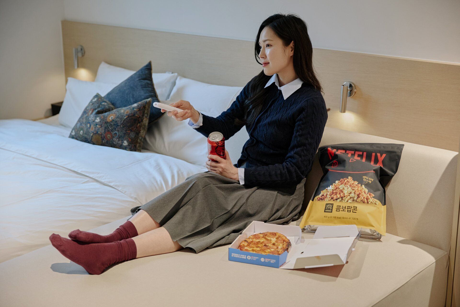 A person sitting on a bed with a pizza box, a can, and a bag of snacks.