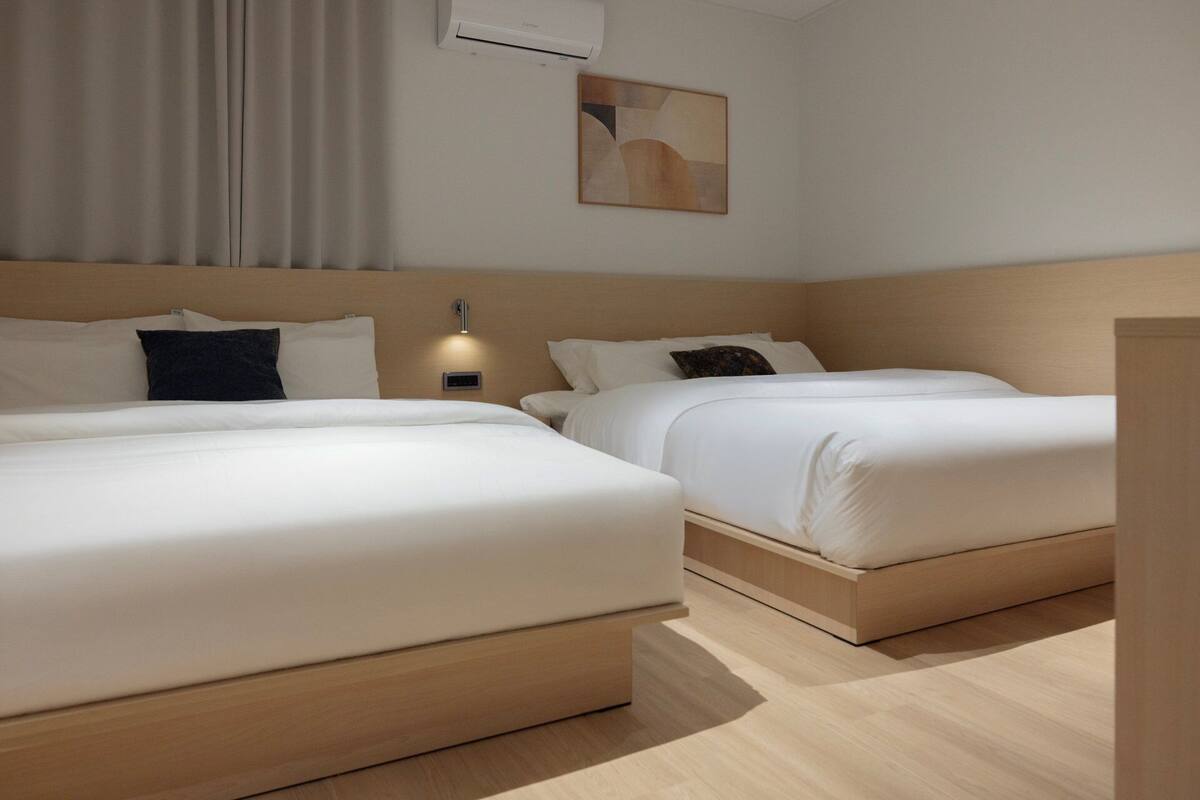 A hotel room with two beds, a headboard, an air conditioning unit, a wall-mounted light, and a painting.