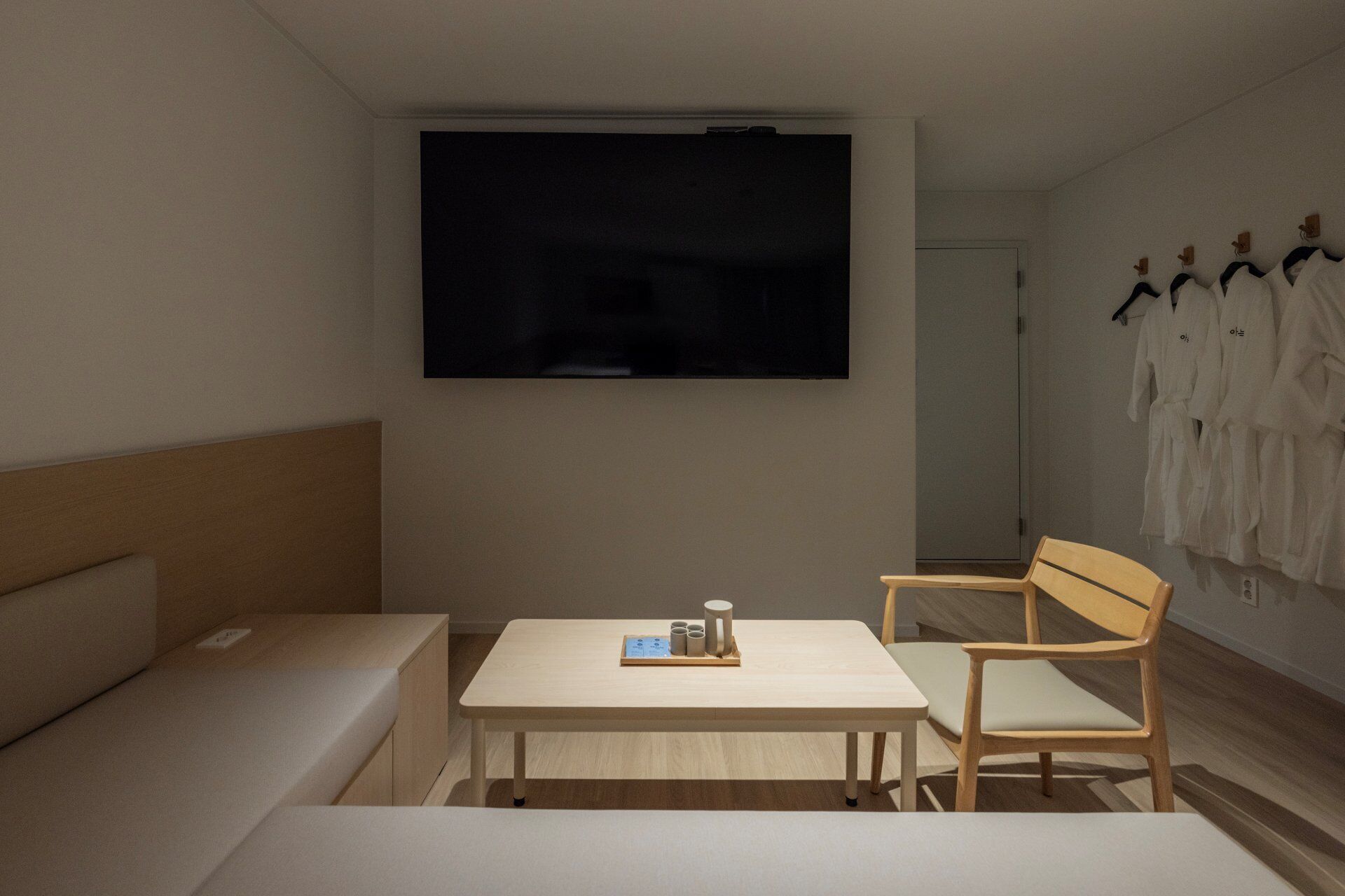 Quarto de hotel com cama, uma mesinha com xícaras, uma cadeira, televisão e um cabideiro com roupões.