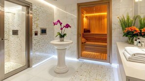 Une salle de bain moderne avec un mur en marbre, un sauna en bois, un lavabo sur pied agrémenté d’orchidées et un meuble-lavabo blanc avec des serviettes.