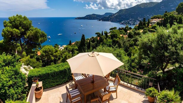 Une terrasse avec une table et des chaises, offrant une vue sur un paysage côtier avec des bateaux et des collines.
