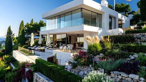 Une maison moderne à deux étages, dotée de grandes fenêtres en verre, d’un espace extérieur aménagé pour se détendre et d’un jardin paysager.