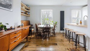 A kitchen with a wooden floor, a dining table with chairs, bar stools, a window, and shelves with various items. - Serene 1BD by London Fields and Victoria Park (Greater London)