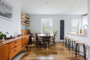 A kitchen with a wooden floor, a dining table with chairs, bar stools, a window, and shelves with various items. - Serene 1BD by London Fields and Victoria Park (Greater London)