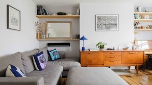 A living room with a sofa, ottoman, wooden cabinet, and shelves with books and decorative items. - Serene 1BD by London Fields and Victoria Park (Greater London)