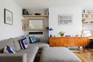 A living room with a sofa, ottoman, wooden cabinet, and shelves with books and decorative items. - Serene 1BD by London Fields and Victoria Park (Greater London)