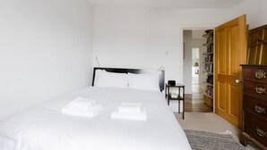 A neatly made bed with white linens and towels, a wooden dresser with drawers, and a bookshelf in a room with white walls and a wooden floor. - Serene 1BD by London Fields and Victoria Park (Greater London)