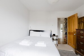 A neatly made bed with white linens and towels, a wooden dresser with drawers, and a bookshelf in a room with white walls and a wooden floor. - Serene 1BD by London Fields and Victoria Park (Greater London)