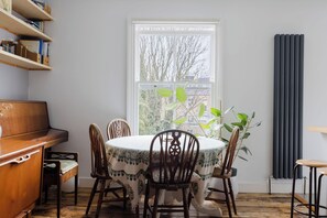 A dining area with a table set for four, surrounded by wooden chairs, a piano, and a bookshelf. - Serene 1BD by London Fields and Victoria Park (Greater London)