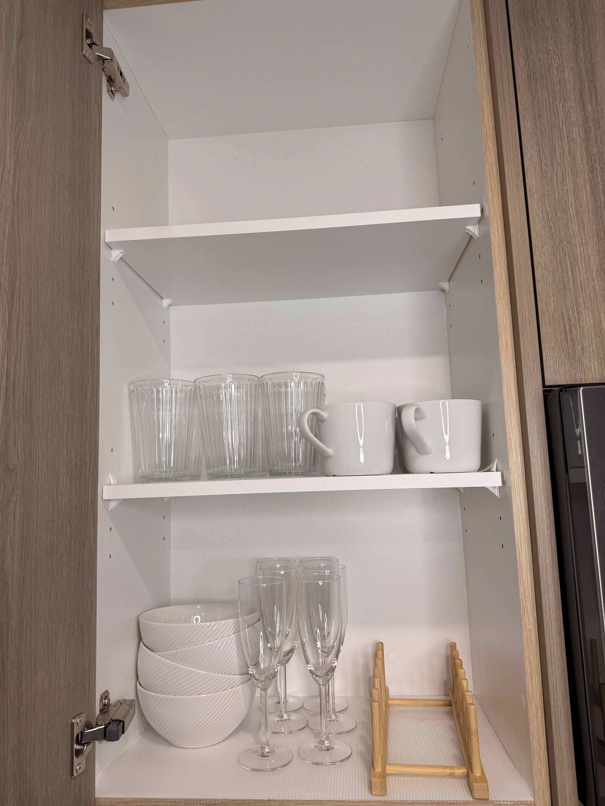 Un mueble de cocina con estantes que contienen vasos de vidrio, una taza blanca y una tabla de cortar de madera.