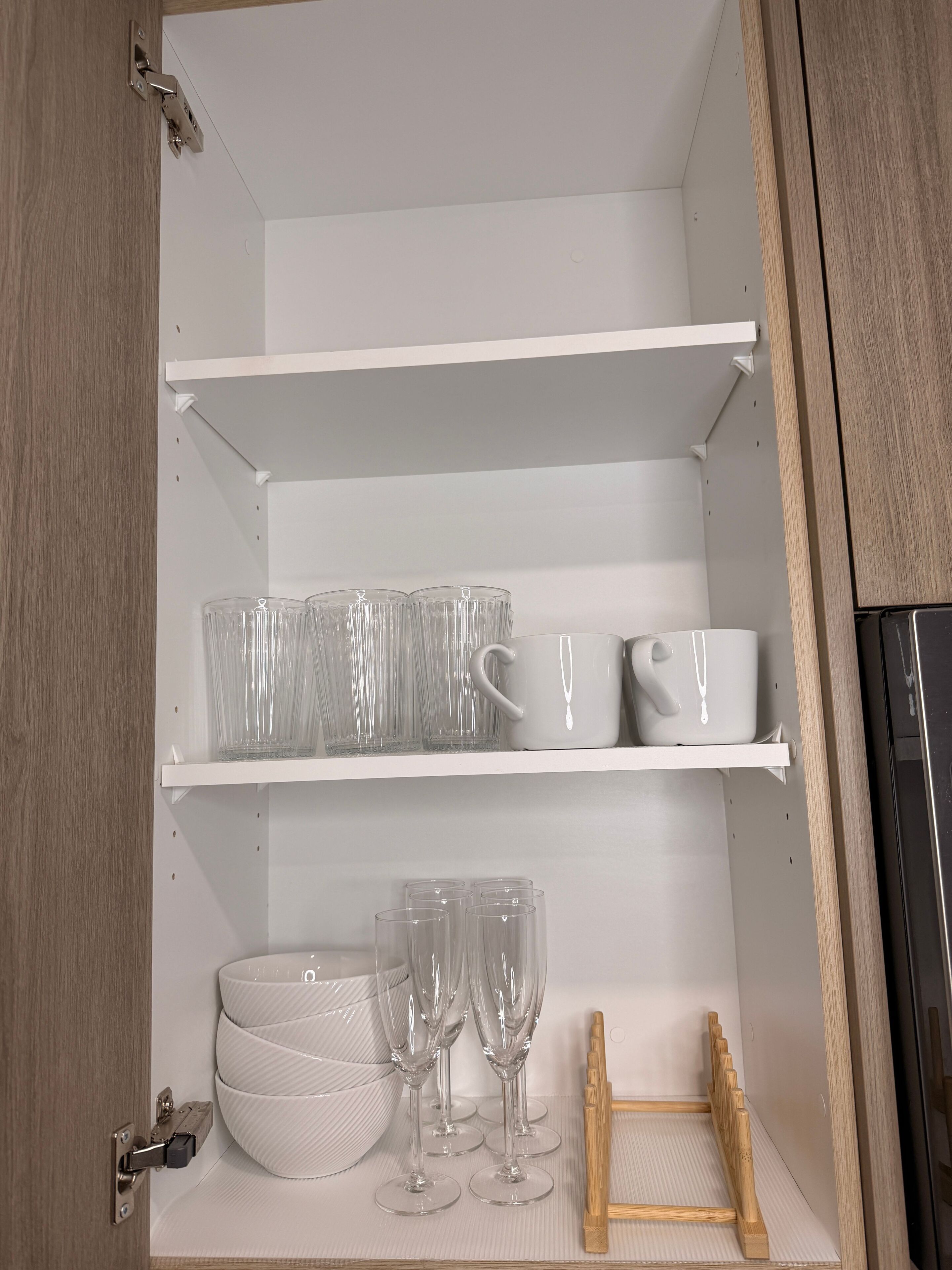 A kitchen cabinet with shelves holding clear glasses, a white mug, and a wooden tray.