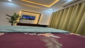 Una habitación de hotel con una cama, un televisor montado en la pared, una planta en maceta y una ventana con cortinas.