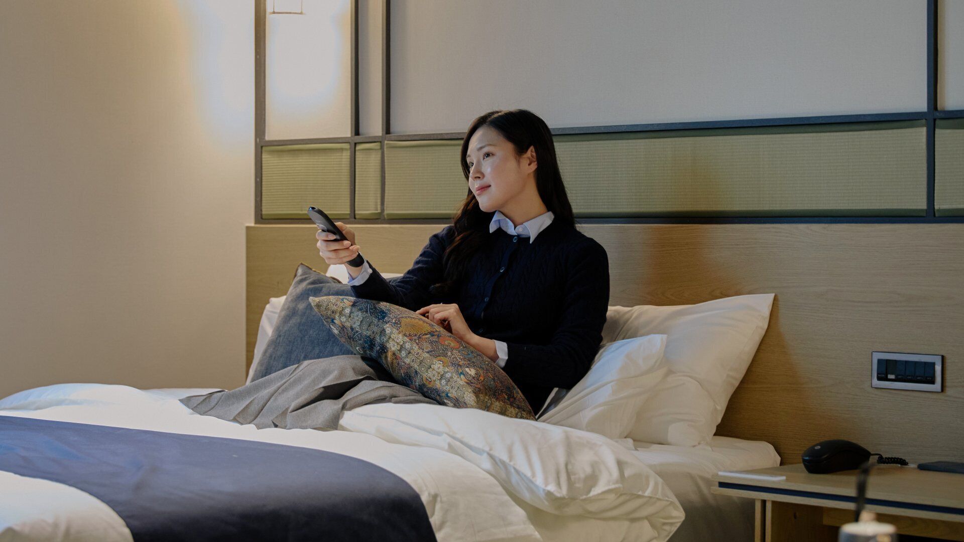 Een hotelkamer met een bed, een nachtkastje met een lamp en een bedieningspaneel aan de muur.