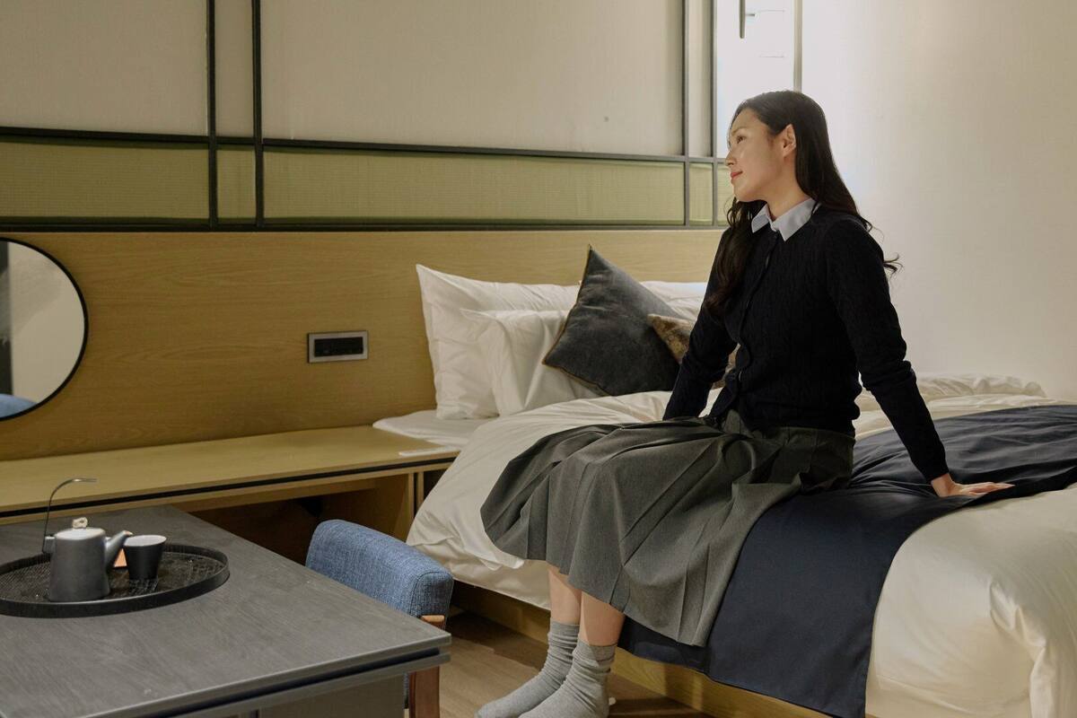 Habitación de hotel con cama, mesita de noche con tetera y taza, una silla y un espejo en la pared.