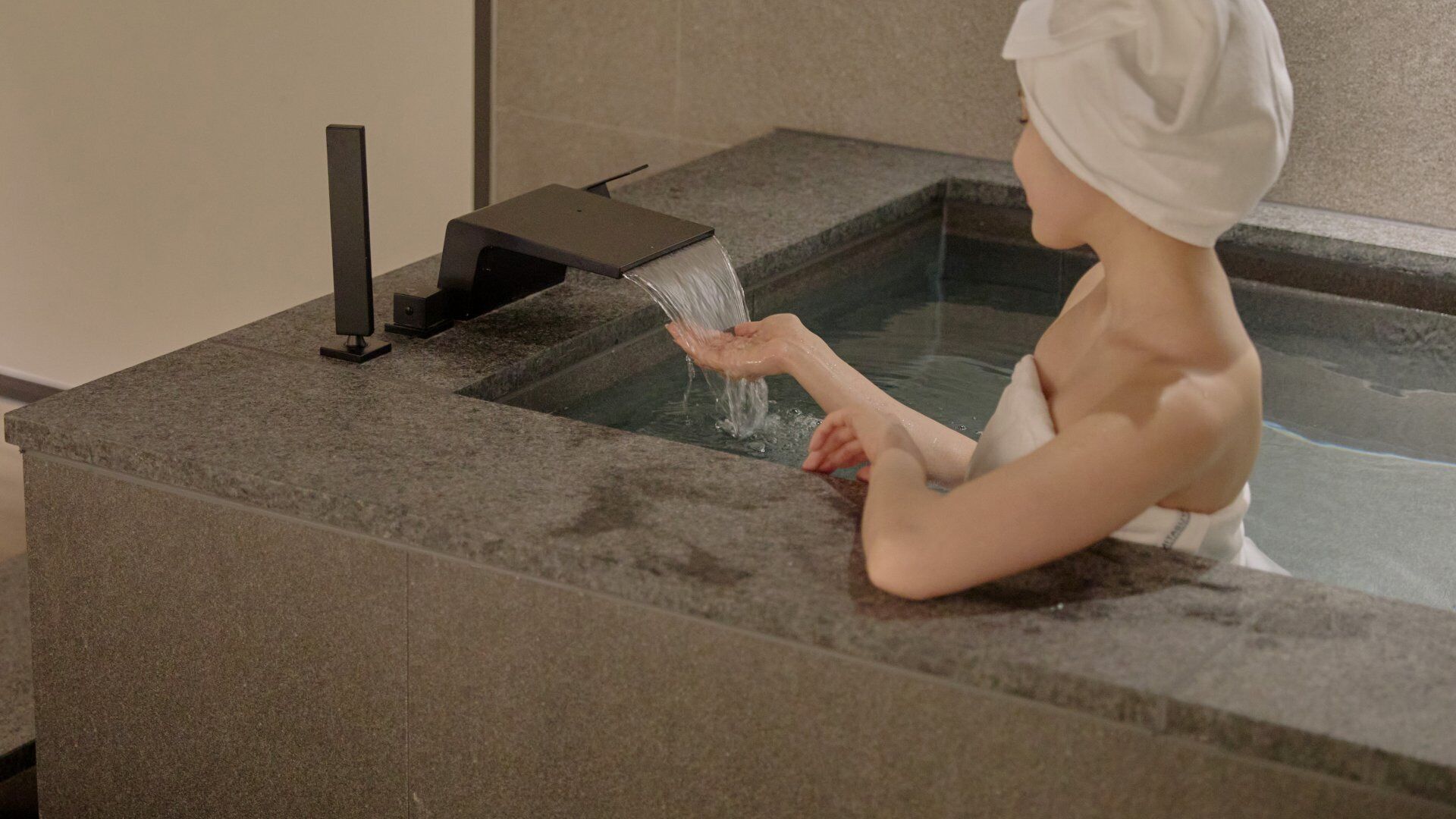 A person in a stone bathtub with a faucet, surrounded by stone walls.