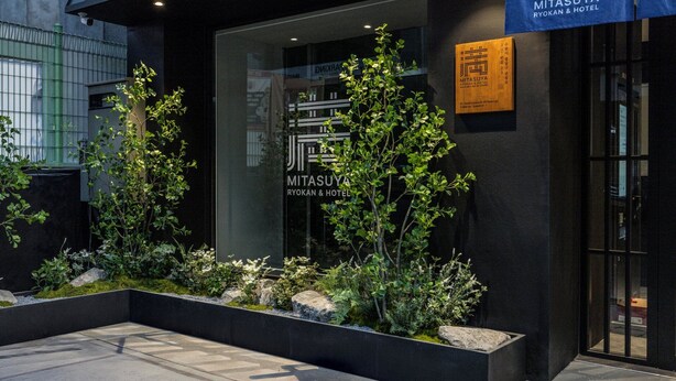 Una entrada de hotel moderna con fachada de vidrio, señalización de 'MITSASUYA RYOKAN & HOTEL' y un jardín cuidado con plantas y rocas.