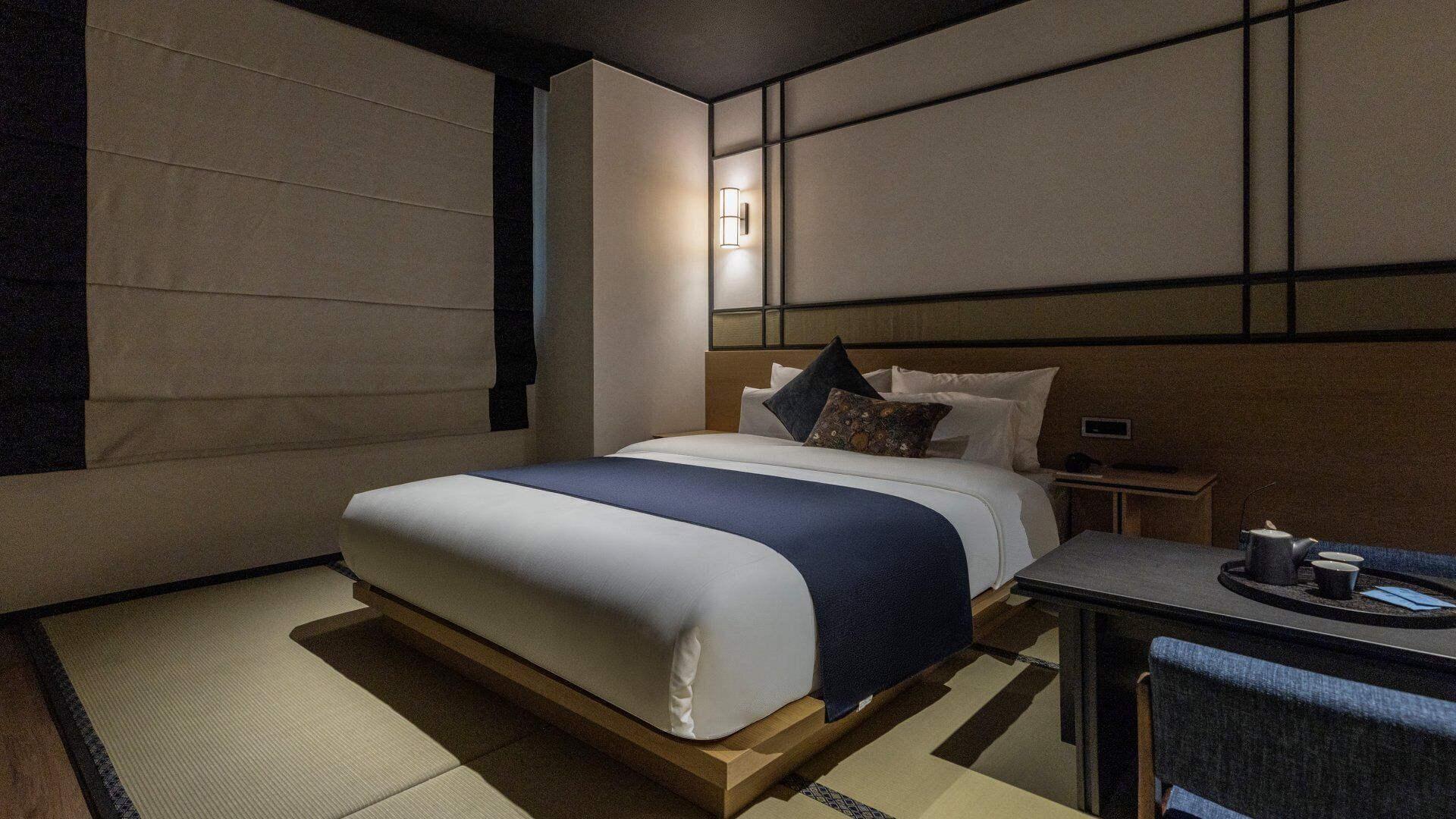 A modern hotel room with a large bed, bedside table, and a small desk with a chair.