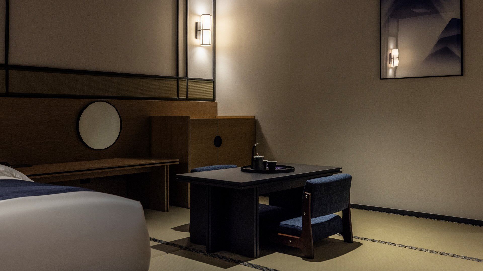 A hotel room with a bed, a desk with a chair, a round mirror, and two wall-mounted lights.