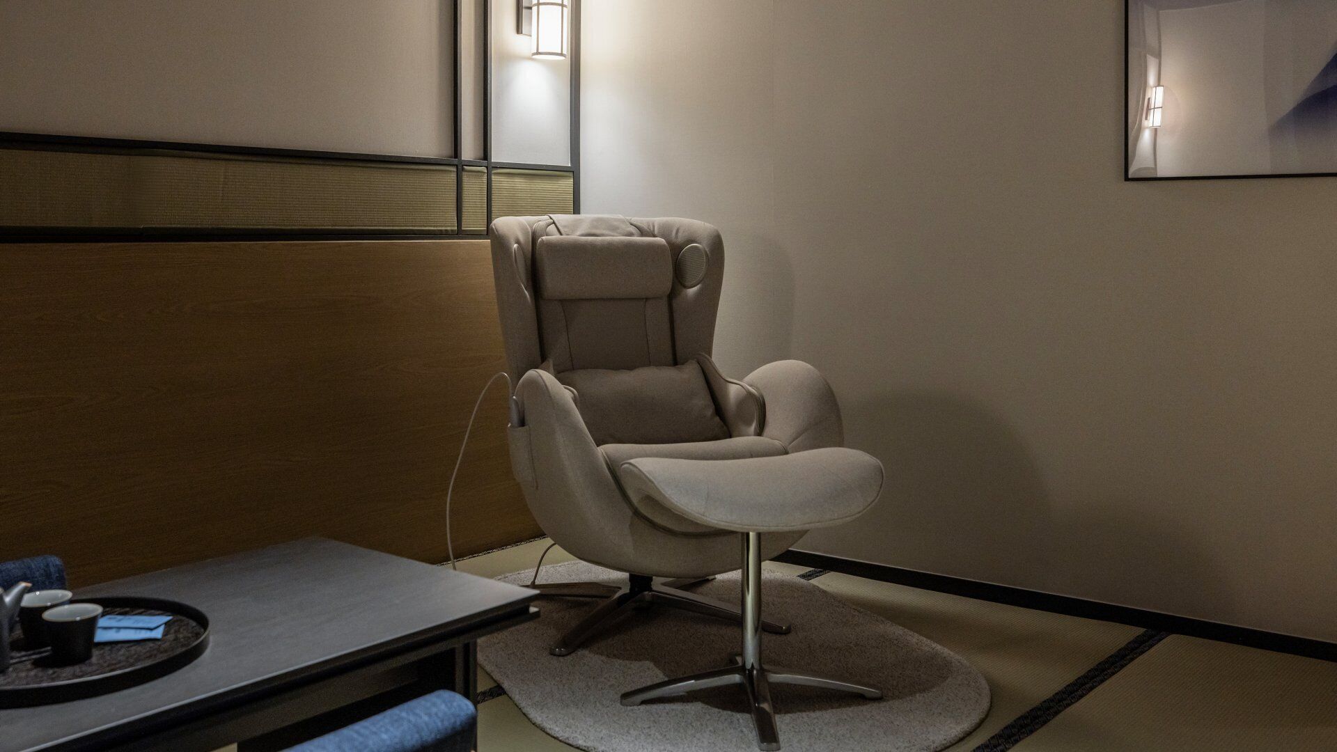 A room with a massage chair, a small table with a tray, and a wall-mounted light.