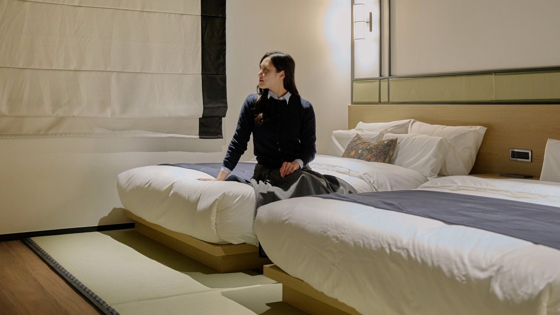 A hotel room with two beds, a large window with curtains, a wooden headboard, and a bedside table with a lamp.