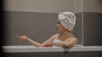 A person in a bathtub with a towel wrapped around their head, set against a tiled wall.