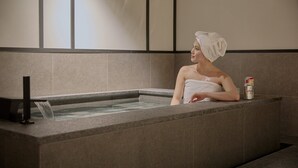 Una persona en una bañera con una toalla en la cabeza, una lata de bebida y una fuente de agua.
