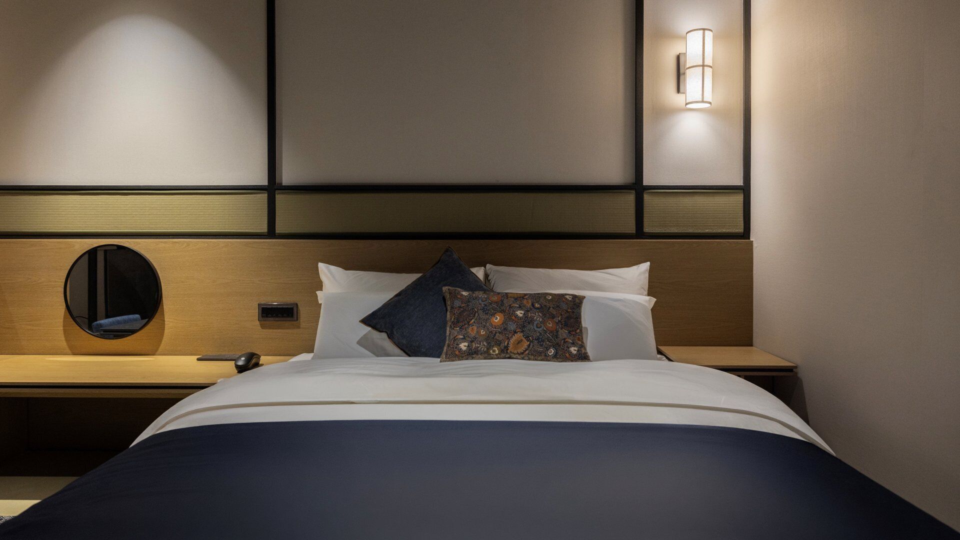 A hotel room with a bed, a bedside table with a lamp, a round mirror, and a control panel.