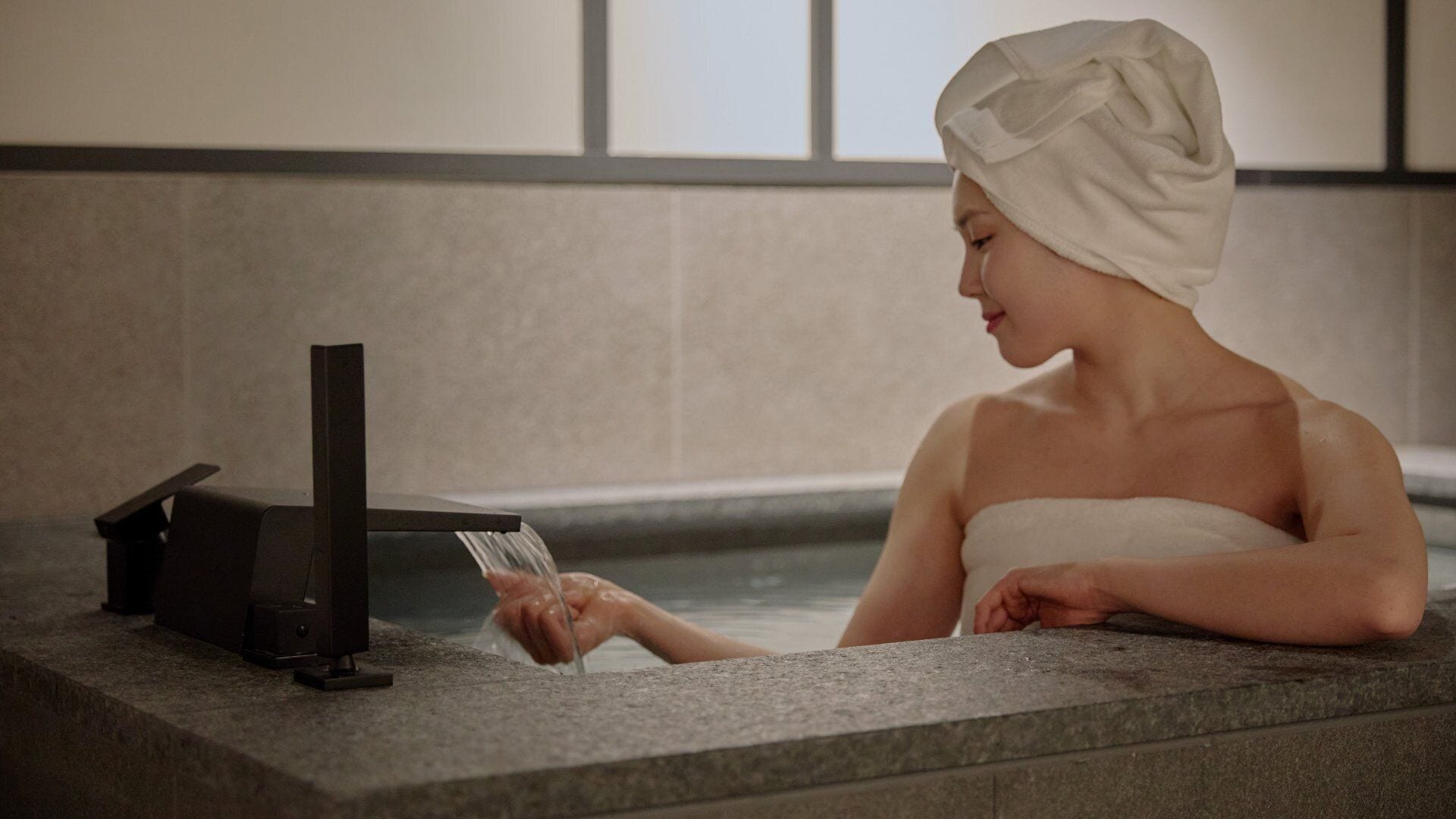 A person in a bathtub with a towel on their head, a water faucet, and a control panel.