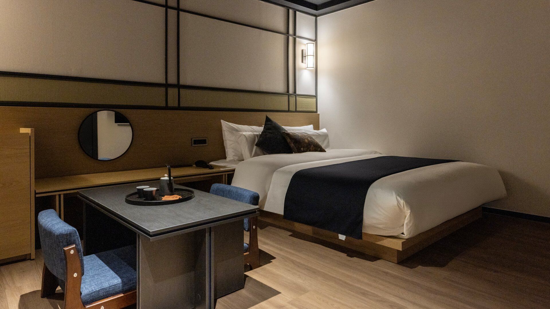 Une chambre d’hôtel comprenant un lit, un bureau avec une chaise, un miroir rond et une table de chevet.
