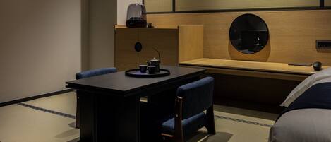 Habitación de hotel con un escritorio de madera oscura, dos sillas azules, una mesita con un hervidor y tazas, un espejo redondo y una cama con almohada blanca.