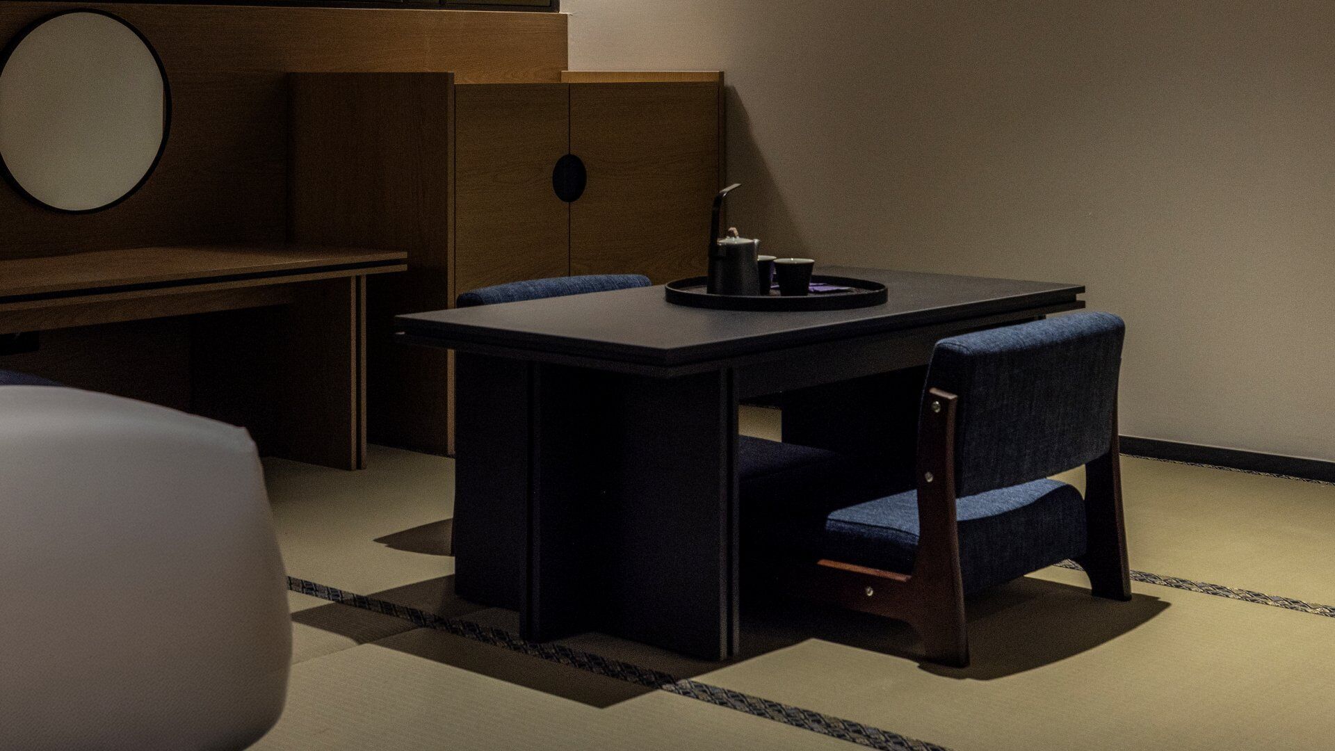 Une pièce avec une table en bois foncé, deux chaises bleues recouvertes de tissu, un miroir rond et un petit objet noir posé sur la table.