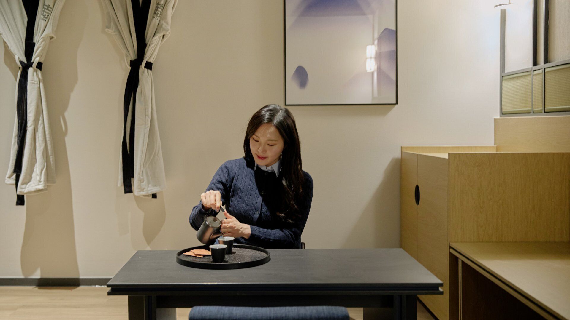 A person sitting at a table with a tray of food, a black table, and wooden furniture in a room with a framed picture on the wall.