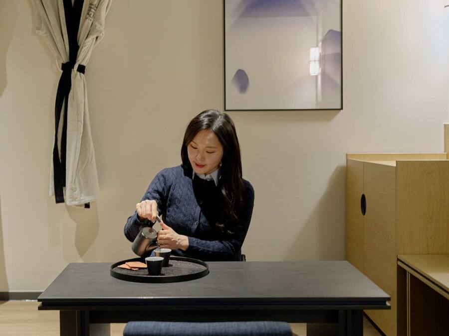 A person sitting at a table with a tray of food, a black table, and wooden furniture in a room with a framed picture on the wall.
