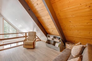 Ein Loft-Zimmer mit hoher Decke, Holzbalken, einem gepolsterten Sessel, einer Couch, einem Bücherregal und einem großen Fenster.