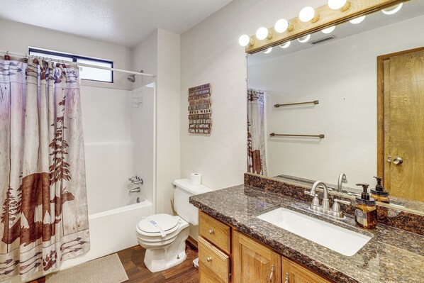 A bathroom with a shower curtain, toilet, sink, vanity, and a mirror with lights. - Bear Canyon Getaway | Hot Tub & Game Loft (Big Bear Lake)